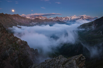 Mountain peaks emerging from the fog at the blue hour while the moon rises, Stelvio National Park, Ponte di Legno, Lombardy, Italy