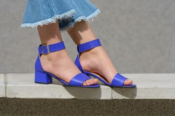 Women's legs in blue denim jeans and sandals in the city street. Trendy elegant casual outfit. Details of everyday summer look. - obrazy, fototapety, plakaty