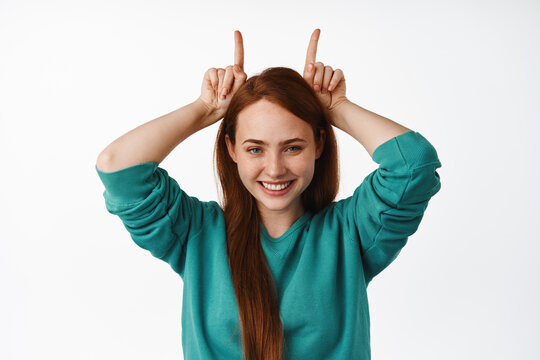Little party never kill nobody. Funny redhead woman looks happy, shows bull horns devil gesture and playing, smiling cute, standing against white background