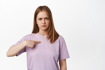 Image of young upset woman pointing at herself and furrow eyebrows from upset, unfair situation,...