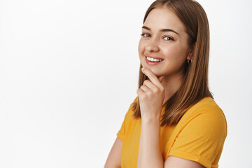 Fototapeta premium Beauty and people concept. Close up portrait of attractive young woman smiling pleased, touch chin and look thoughtful at camera, standing confident against white background