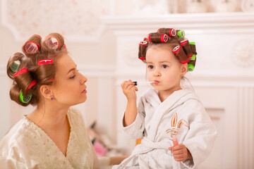 Adorable little girl with her mother in hair curlers apply makeup. Mom teaches daughter to use cosmetics. Beauty day. Girls are such girls