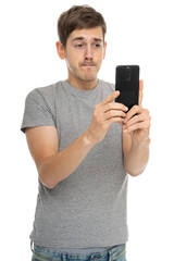 Young handsome tall slim white man with brown hair is unsure in gray shirt with mobile phone isolated on white background