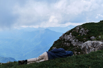 Naklejka premium Traveler was tired in campaign and decided to rest. Male tourist fell asleep in grass on top of mountain against the background of low clouds and snow capped mountains in distance.
