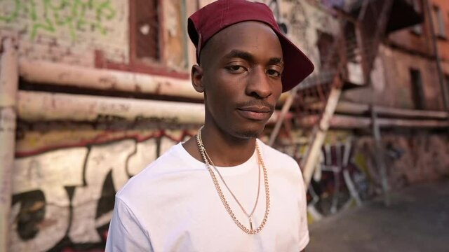 Portrait of a young black guy on the street in the ghetto. Young man calmly looks at the camera against the background of a wall with graffiti on the street. Slow motion