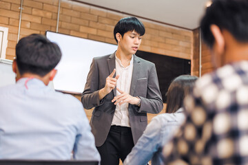 business cooperation : Young asian male coach or speaker make flip chart presentation to diverse businesspeople at meeting in office. Male tutor or trainer present project to diverse colleagues.