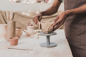 partial view of young african american man sculpting clay pot with spatula on table with equipment...