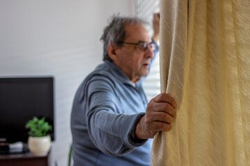 Shot of a thoughtful old man looking out of the window at home. A senior Caucasian man in his 70s at home at a window in the bedroom, opening the curtains to let the sunlight in. Focus on hands.