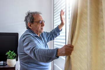 Naklejka na ściany i meble Photo of a senior man is waving from a window in his home. Happy elderly man standing by the window. Old man with eyeglasses standing by the window and looking out with raised hand, waving to someone.
