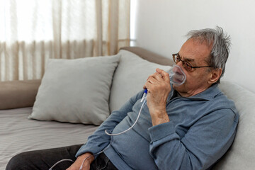 Senior man using medical equipment for inhalation with respiratory mask. Old man with serious pneumonia symptoms caused by Covid-19 virus infection sitting in home during lockdown, wearing oxygen mask