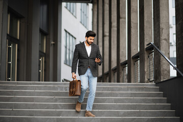 Handsome arabian man dressed in stylish formal clothes standing on stairs with suitcase and smartphone in hands. Modern gadget for work. Business concept.