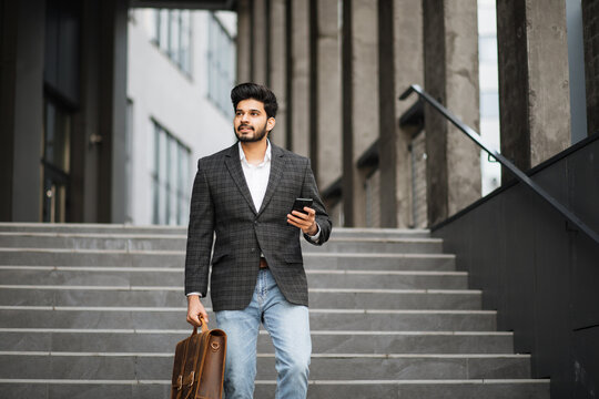 Handsome arabian man dressed in stylish formal clothes standing on stairs with suitcase and smartphone in hands. Modern gadget for work. Business concept.
