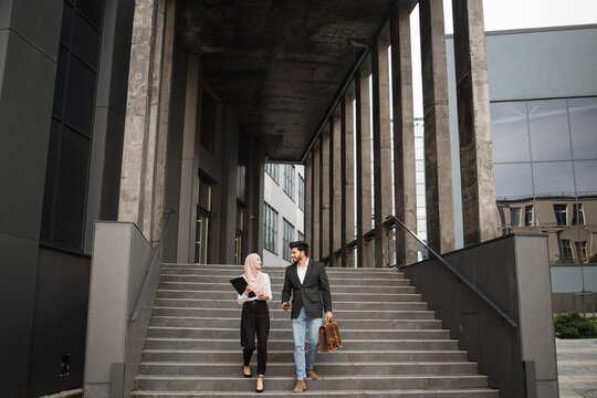 Full length portrait of muslim woman and man walking together near modern office building an discussing some working moments. Two coworkers with smartphones, clipboard and suitcase outdoors.