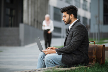 Side view of arabian businessman using modern laptop for remote work while sitting on bench outdoors. Blur background of charming woman in hijab walking and using smartphone.