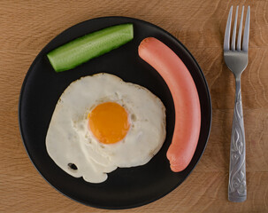 Fried egg, cucumber and sausage on a black plate, which is located in the place with a fork on a wooden board