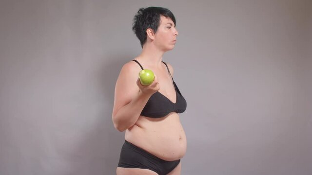 Overweight woman eats an apple. Following a diet, eating fruit. Satisfying appetite with low-calorie food. Hand touches belly fat, fullness awareness.