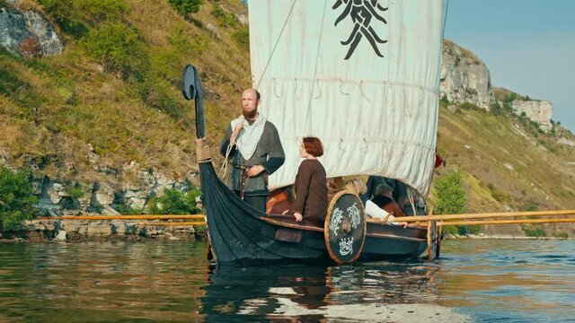 A Viking man Stands Aboard the Drakkar Dressed in Armor and Looks into the Distance. Vikings Sail on an Old Ship with a Raised Sail Along a Calm River Against the Backdrop of a Rocky Coastline.
