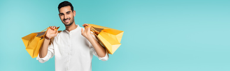 Positive arabian man holding yellow shopping bags isolated on blue, banner