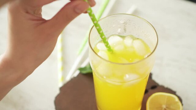 Girl without face mixes an orange drink with light green cocktail tube and drinks refreshing drink. Glass of orange soda with ice and straw on wooden slice. There are several paper tubes on white tabl