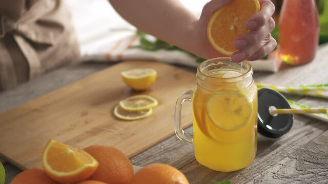 Woman's hand crushes orange juice on wooden kitchen table. Orange juice with soda on a wooden table. Soft drink with citrus slices, orange soda in a glass with a Mason handle. Delicious orange sparkli