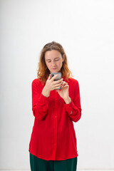 young concentrated caucasian redheaded pretty woman with long hair in red shirt isolated on white background holds smartphone in hands, chats on web. Technology, communication, using mobile phone