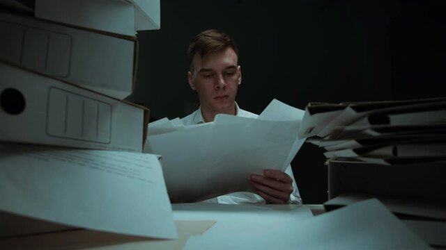 Tired man with pile of folders working in office, employee works with documents in evening having headache, getting new task, exhausted worker holding his head. Bad mood, fatigue, lots of work. 