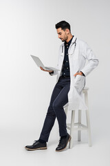 Side view of arabian doctor using laptop near chair on grey background