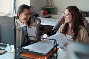 two young latina business women talking, laughing and working together in the office, using documents and office supplies, concept of teamwork.