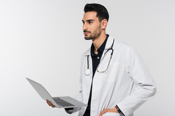 Young muslim doctor holding laptop and looking away isolated on grey