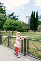 Little girl in a panama hat stands near a metal fence in a green park