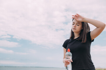 A girl of Caucasian appearance, after jogging, wipes sweat from her forehead and holds a bottle of...