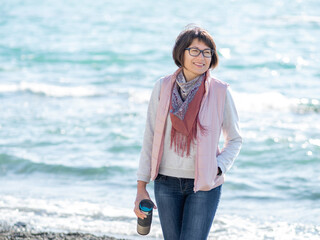Smiling woman walking on seaside with metal travel mug. Vacation on ocean coast. Turquoise water...