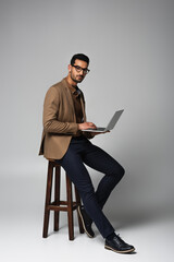 Arabian businessman using laptop and looking at camera on chair on grey background