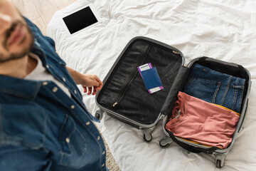 High angle view of blurred man standing near suitcase with passport and digital tablet on bed