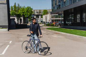 Fototapeta na wymiar Young muslim manager in jacket holding coffee and bicycle on urban street