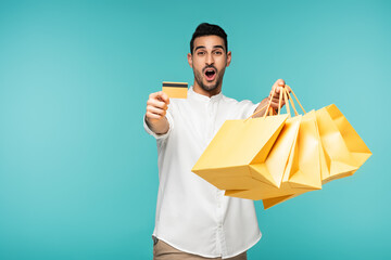 Amazed arabian man holding credit card and shopping bags isolated on blue