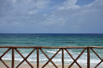Seashore with white clouds in the blue sky, with the top view of a  thin wooden railing.