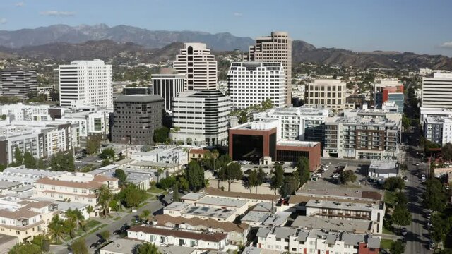 Aerial Panning Sunny Downtown City Buildings And Urban Skyline With Bright Sunlight, White Towers, And Rugged California Mountains On The Horizon - Glendale, California