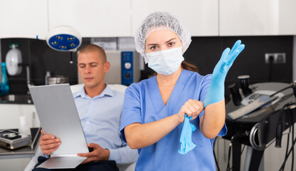 Confident woman beautician putting on gloves before consultation at aesthetic clinic, man sitting on couch and looking at mirror on background