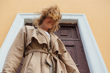 Young millennial woman with wild hair dressed in an autumn coat posing near the door of an old...