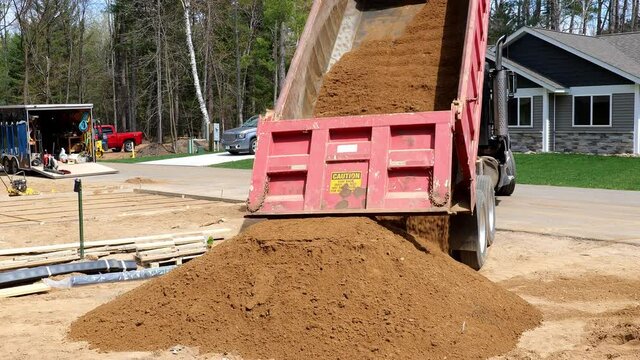 A gravel box tilted up dumping a load of dirt. Red dump truck with black cab at a new home construction site.