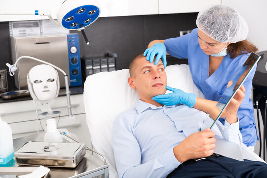 Portrait Of Man Sitting On Couch And Looking At Mirror During Appointment With Doctor At Aesthetic Clinic