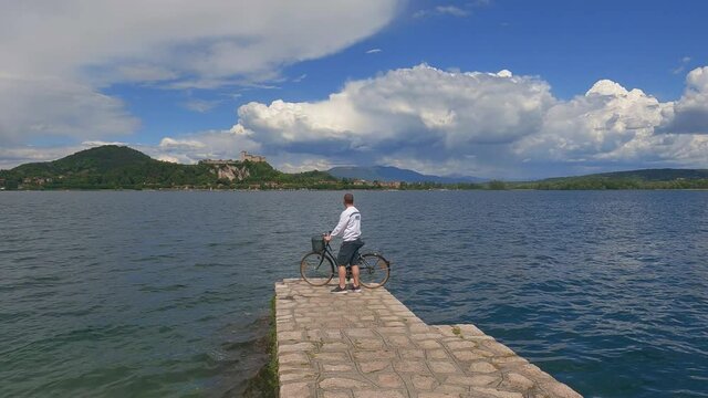 Man with bicycle standing on dock edge of lake Maggiore in Italy, Angera castle on background. Static view