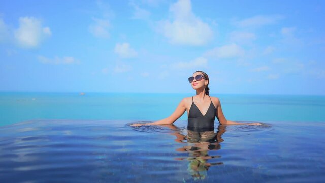 Confident exotic female in a black swimsuit in infinity pool with a tropical sea horizon in background, luxury vacation concept, full frame