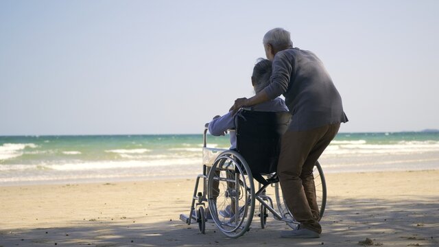 Happy Asian back elderly woman disabled sitting in wheelchair and husband is a wheelchair user on the beach together, summer vacation, Retirement couple concept - Powered by Adobe