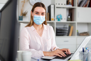 Fototapeta na wymiar Female office worker in protective medical mask is having productive day at work in office