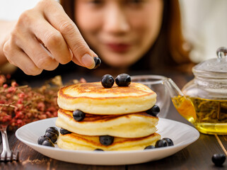 Close up hand. A woman holding hand decorating Pancake by picking up blueberries and blur teapot background. Concept sweet food.