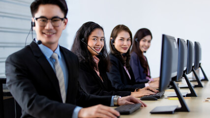 Cheerful Asian man in headset smile with cheerful and happy while working on computer at desk with female coworkers in help desk office. Sevice mind and ready  for marketing support concept