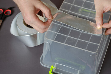 A man measures the length of a piece of aluminum tape over the bottom of a plastic container. The process of repairing a crack in a food container.  View from above from an angle. Selective focus.