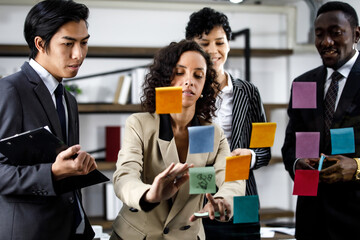 Close up to African beautiful smart professional black formal businesswoman thinking and writing creative marketing idea list on colorful sticky notes at mirror surrounded by multiracial colleagues.